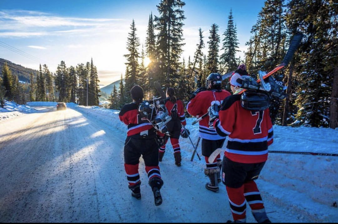 A group of four women in hockey gear walking up a snowy path with sun streaming through the trees in the background.